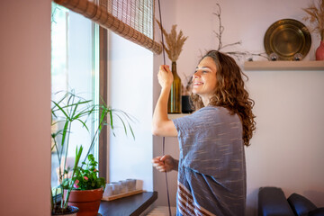 Beautiful happy young woman opening rolled bamboo blinds in country house in summer morning - obrazy, fototapety, plakaty