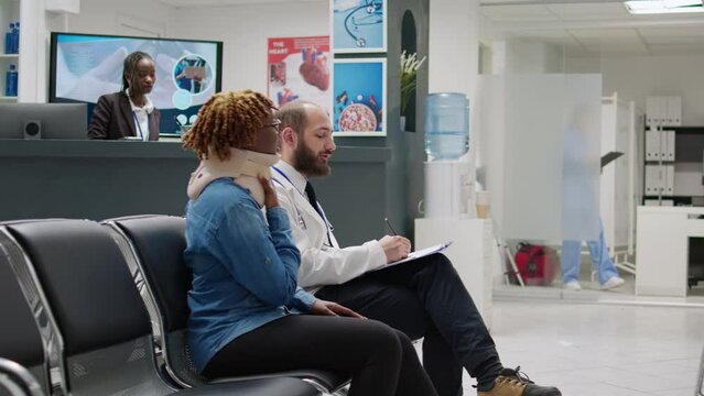 African american woman with cervical collar being hurt in pain, talking to doctor in waiting area at medical facility. Patient with physical injury wearing neck foam brace after accident trauma.
