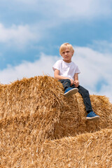 Naklejka na ściany i meble Blond boy of European appearance in jeans and t-shirt sits on haystack on blue sky background.