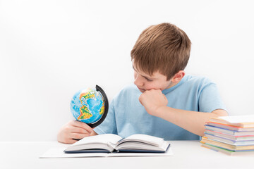 Boy sits at table with globe in hands and thinks about something. Student does homework and carefully scrutinizes globe.