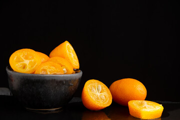 Close-up of wet and cut kumquats, in dark bowl, black table, selective focus, black background, horizontal, with copy space
