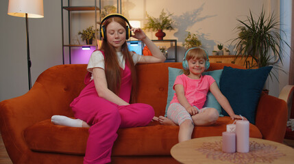 Happy relaxed overjoyed two girls friends siblings in wireless headphones relaxing at home couch choosing listening dancing favorite energetic disco music. Female child kids family couple. Friendship