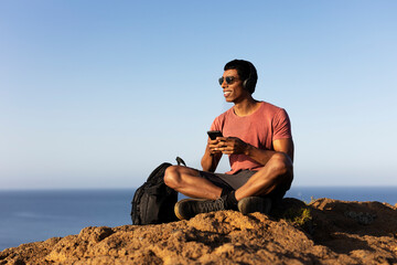 Young man sitting on the stone listening the music. Tourist man on the top of the mountain..