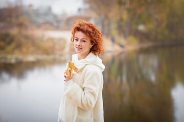 Attractive stylish woman dressed in white warm coat holding yellow leaves walking near river. Autumn mood concept