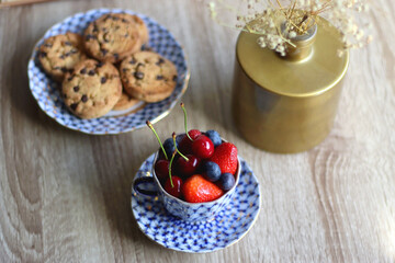 Plate of chocolate chip cookies, cup filled with strawberries, blueberries and cherries, open book and vase with gypsophila flowers on the table. Selective focus.