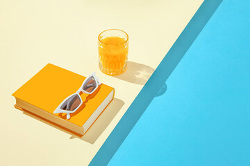 Creative summer scene with orange book, stylish white sunglasses and crystal glass on sunny blue...