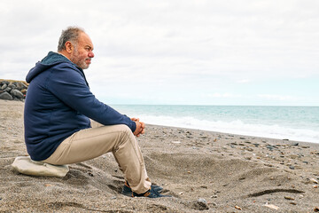 Side view of thoughtful bearded mature man sitting on pebble winter beach and contemplating blue...