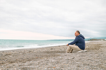 Side view of thoughtful bearded mature man sitting on pebble winter beach and contemplating blue sea. Concept of leisure activities,tourism, lifestyle e nature.