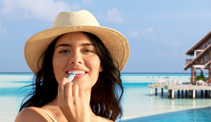 beauty, cosmetics and summer holidays concept - portrait of happy smiling young woman in bikini swimsuit and straw hat applying lip balm over tropical beach background in french polynesia