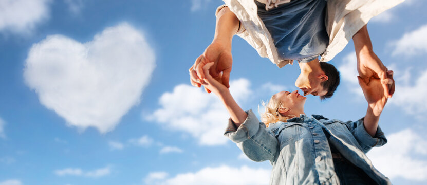 summer holidays, love and people concept - happy young couple holding hands under blue sky with cloud in shape of heart on background, from below