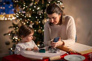 family, cooking and winter holidays concept - happy mother and baby daughter with smartphone making gingerbread cookies at home on christmas