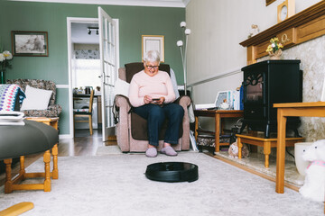Senior woman using a robot vacuum cleaner through phone settings while sitting on armchair at home. Mature older lady holding mobile phone and control cleaning. Smart home. Seniors and technology - obrazy, fototapety, plakaty
