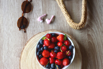 Round straw bag, brown sunglasses, pink earphones and bowl of berries on wooden table. Flat lay.