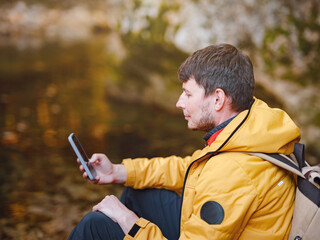 Adventure and active lifestyle in nature. Tourist hiking in forest. Caucasian man in yellow jacket walks in woods. using navigation application on smartphone, social networks, taking pictures