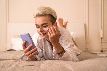 Blonde girl with under eye patches scrolling social media while laying at the bed after shower