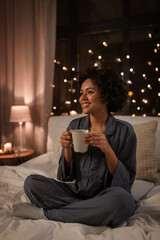 people, bedtime and rest concept - happy smiling woman in pajamas with coffee sitting in bed at night