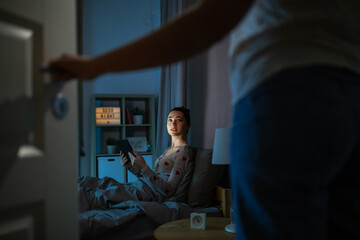 technology, bedtime and people concept - teenage girl with tablet pc computer sitting in bed at home at night and mother entering room