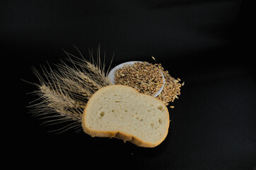 Grains of wheat on a plate next to white bread and ears of wheat on a black background