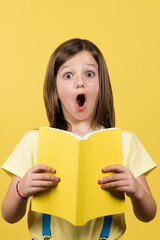 Portrait of surprised little girl holding book isolated on yellow background.
