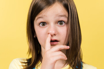 Little girl drilling the nose with finger to remove mucus isolated on yellow background. Funny child portrait.