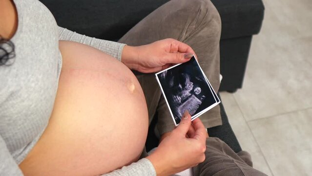 A pregnant woman in comfortable clothes sits on the floor and looks at an ultrasound photo of her baby. Twins. Top view