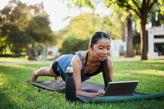 Sporty girl working out on a mat on the grass in the park while using a tablet computer