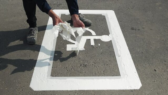 Close-up of manual utility male worker peeling off white tape from freshly painted white disabled car parking lot sign, disability symbol, wheelchair painting on road asphalt surface, summer daytime.