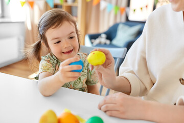 holidays, family and people concept - happy little baby daughter and mother tapping colored easter eggs at home