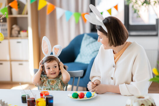 holidays, family and people concept - happy mother and little baby daughter in bunny ears dyeing easter eggs at home