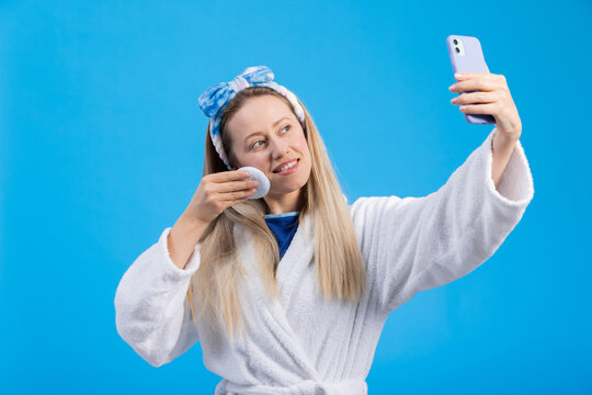 Influencer girl takes photos of herself and records a video during the morning facial care. Young woman in a bathrobe on a blue colored studio background.