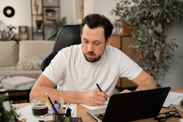 A busy man works at workplace. He holds pen in hand and writes down the main points of report. On the table is open laptop, cup of coffee and stationery. The man is focused on his work.