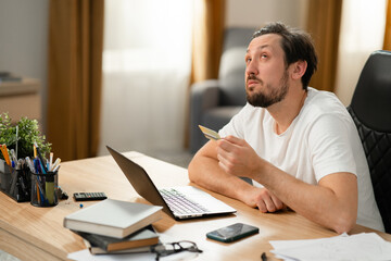 Fototapeta premium Young brunette with beard ponders new purchase online. In his hands he holds bank card in hand. He thinks it's better to buy. On the table lies open laptop book calculator phone.