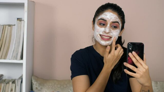 A young Indian college student with a face mask - checking herself on a mobile camera  skin care  skin routine. A cheerful Indian female with a face pack posing while taking pictures on her smartph...