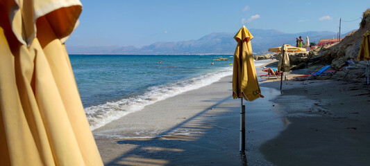 Beautiful view of a beach with folded yellow sun umbrellas, a lapping wave, and mountains in the...