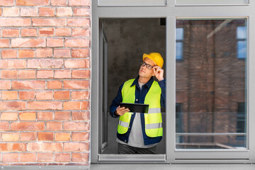 architecture, construction business and building concept - male architect in helmet and safety west with tablet pc computer in window