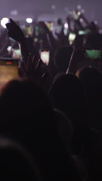 Vertical video of unrecognizable fans dancing at a concert or festival party. Silhouettes of concert crowd in front of bright stage lights