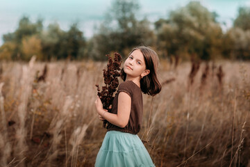 A 10-year-old girl is walking in a field. Dry grass all around. The girl is holding a bouquet. The girl is wearing a brown t-shirt and a blue skirt.