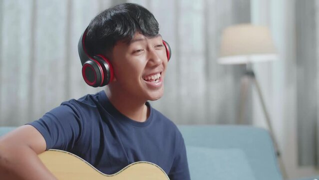 Close Up Of Asian Boy Composer With Notebook On Table Wearing Headphones Singing And Playing Guitar At Home
