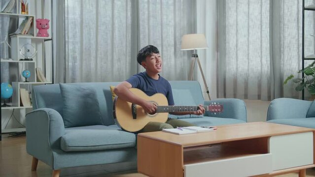 Asian Boy Composer With Notebook On Table Singing And Playing Guitar At Home
