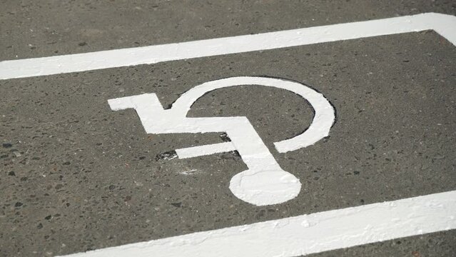 Close-up, circular pan shot of freshly painted white disabled car parking lot sign, disability symbol, man on wheelchair, painting on road asphalt surface, summer daytime.