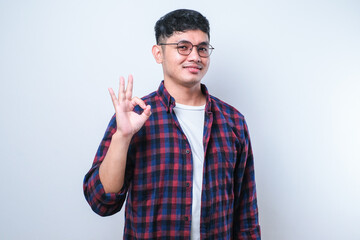 Young handsome Asian man wearing casual shirt and glassessmiling, doing ok sign, thumb up with fingers, excellent sign