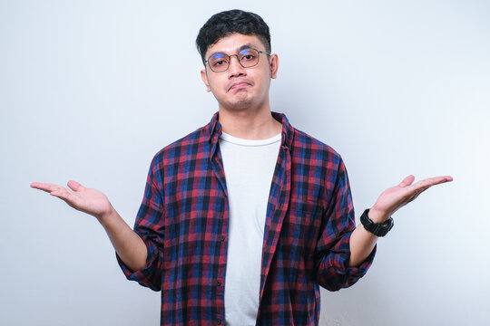 Young Handsome Asian Man Wearing Casual Shirt And Glasses Cueless And Confused Expression With Arms And Hands Raised.