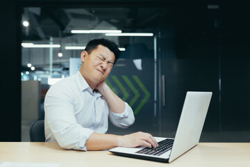 Naklejka na ściany i meble Overtired asian worker in office, having severe neck pain, massaging with hand, businessman working in office with laptop