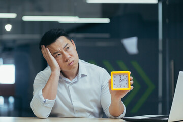 Tired and overworked asian businessman working late in office, holding clock, waiting for decision result