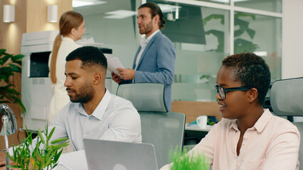 Fototapeta premium A black woman with short hair and glasses is talking to her colleague that s mixed race with a beard in a large lit office.