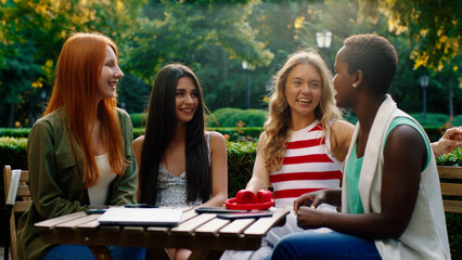 A beautiful and very close friend group of girls are at a park, sitting at a coffee table with all...