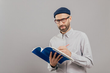 Horizontal shot of concentrated male copywriter writes ideas for new strategy, holds blue notepad,...