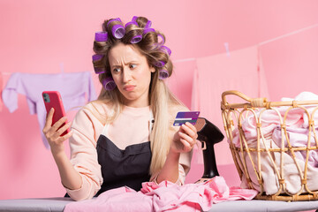 A woman with hair rollers on her head does housekeeping irons clothes hangs laundry on a string. Girl orders cleaning products on phone, purchases pay with bank card using app.
