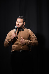 indian stand up comedian grimacing and showing thumb up while telling jokes into microphone on black.