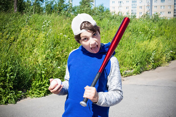 Boy with baseball bat and ball against green grass, mouth open and tongue out, outdoors on sunny day